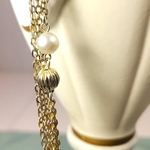 Puede incluir: Primer plano de un collar dorado con m&uacute;ltiples cadenas. El collar presenta una perla y una cuenta acanalada de color dorado. El fondo es un objeto decorativo borroso de color blanquecino.