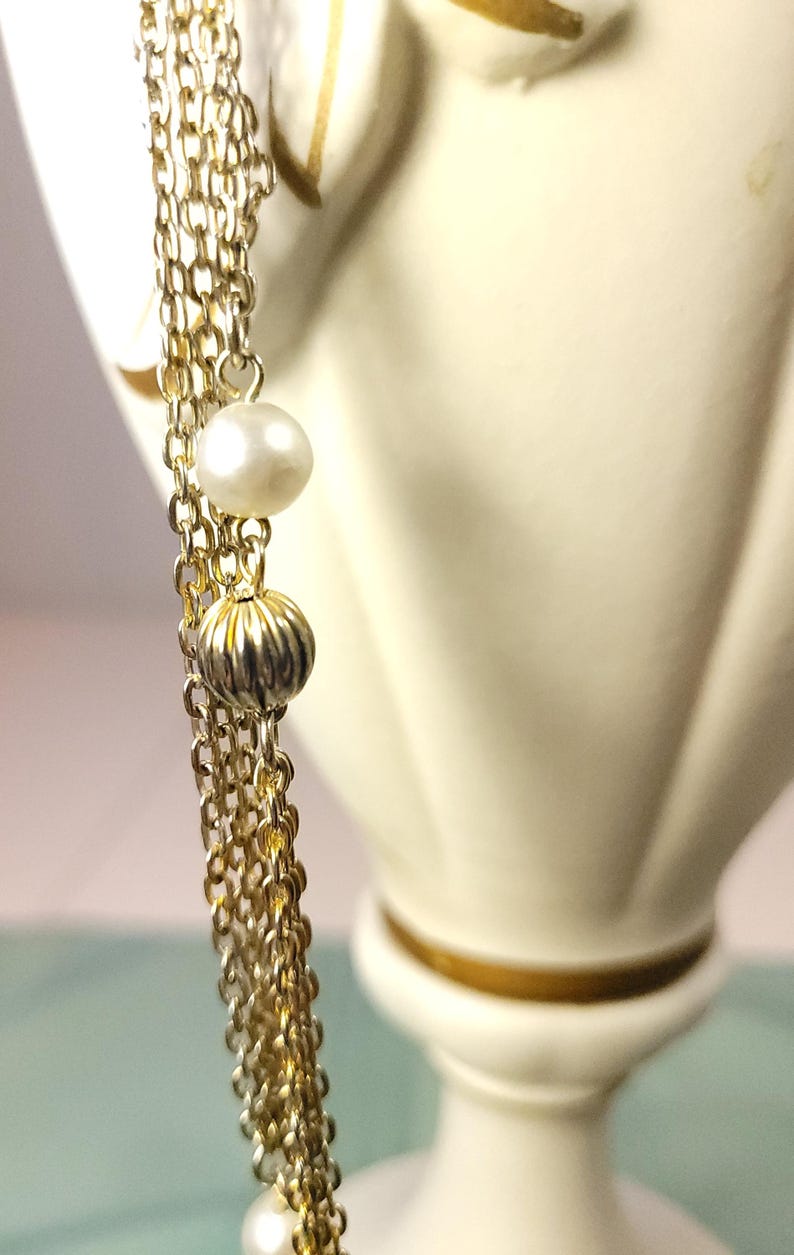 Puede incluir: Primer plano de un collar dorado con m&uacute;ltiples cadenas. El collar presenta una perla y una cuenta acanalada de color dorado. El fondo es un objeto decorativo borroso de color blanquecino.