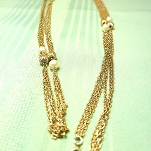 Puede incluir: Collar dorado con m&uacute;ltiples hilos y detalles de perlas. El collar presenta delicadas cadenas y peque&ntilde;as perlas redondas agrupadas a intervalos. El cierre es de dise&ntilde;o simple y circular. El collar se muestra sobre un fondo verde claro.