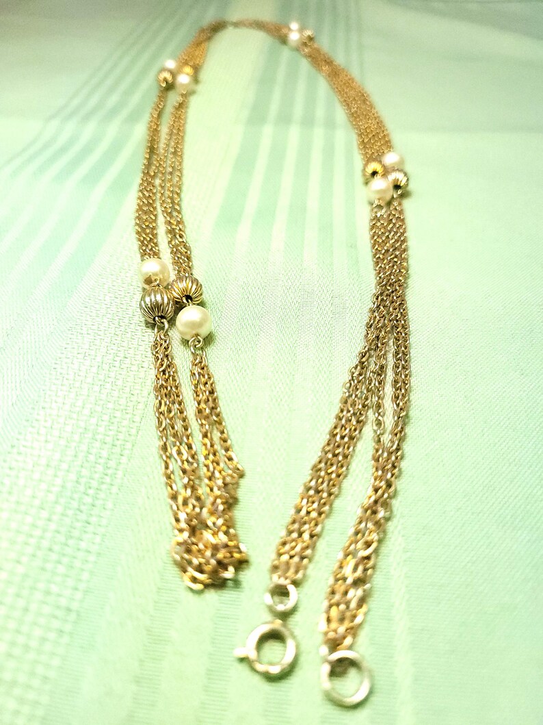 Puede incluir: Collar dorado con m&uacute;ltiples hilos y detalles de perlas. El collar presenta delicadas cadenas y peque&ntilde;as perlas redondas agrupadas a intervalos. El cierre es de dise&ntilde;o simple y circular. El collar se muestra sobre un fondo verde claro.