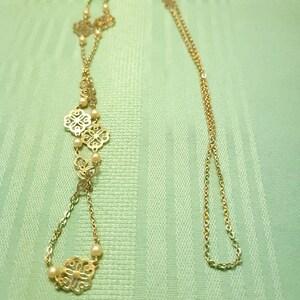 May include: A gold-coloured necklace with intricate floral pendants and small pearls. The necklace has a long chain and a drop pendant design. The design is ideal for adding a touch of elegance to any outfit.