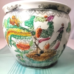 Puede incluir: Una maceta de cerámica blanca con un diseño detallado pintado a mano. El diseño presenta un pavo real colorido con plumas naranjas, verdes y amarillas, rodeado de flores y follaje. El borde tiene un borde decorativo en blanco y negro.