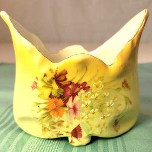 Puede incluir: Un jarrón de cerámica amarilla con forma de tulipán acampanado. El jarrón está decorado con un diseño floral en tonos rojos, naranjas y blancos. El borde está adornado con oro. El jarrón tiene un pie pequeño.