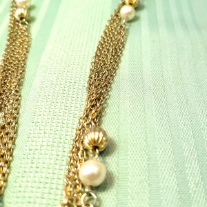 Puede incluir: Primer plano de un collar dorado con m&uacute;ltiples hilos. El collar presenta peque&ntilde;as perlas redondas y cuentas doradas decorativas. El fondo es una tela verde claro, que proporciona un tel&oacute;n de fondo contrastante para la joya.