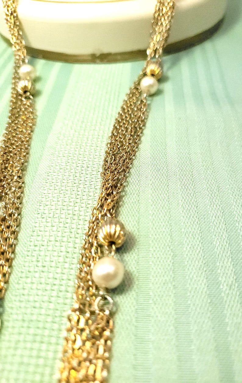 Puede incluir: Primer plano de un collar dorado con m&uacute;ltiples hilos. El collar presenta peque&ntilde;as perlas redondas y cuentas doradas decorativas. El fondo es una tela verde claro, que proporciona un tel&oacute;n de fondo contrastante para la joya.