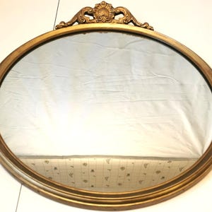 May include: Oval gold-framed mirror with decorative detailing at the top. The mirror's frame has a distressed finish, and the glass reflects the surroundings. A decorative piece for home decor.