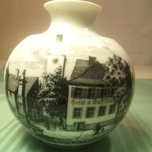May include: White ceramic vase with a black and white illustration of a European village scene. The vase features a round body and a narrow neck. The illustration includes buildings, trees, and the text "Hotel u. Gasthof".