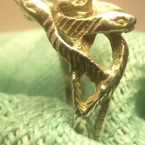 May include: A gold-colored ring with a detailed design featuring a serpent or dragon-like creature. The ring's design is intricate, with textured surfaces and a unique shape. The ring is set against a light green fabric.
