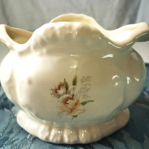 May include: Cream-coloured ceramic bowl with a scalloped rim and base. The bowl features a floral design with peach and white flowers and green leaves. The bowl has a rounded shape and is likely for decorative use.