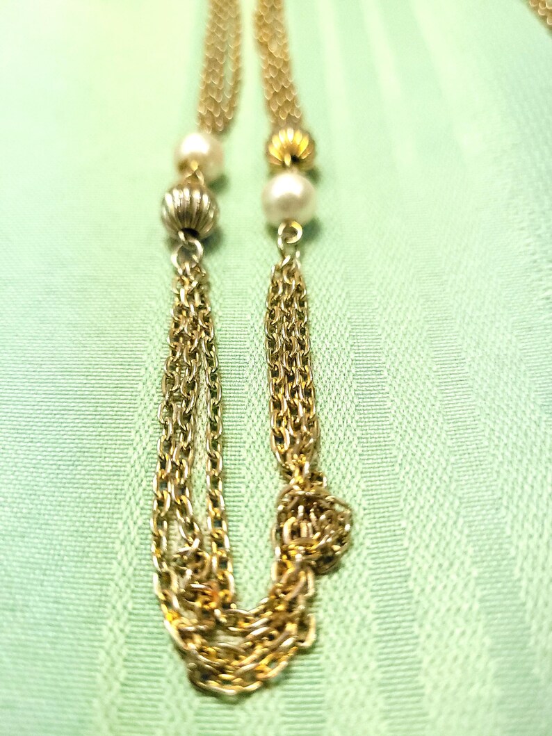 Puede incluir: Un collar dorado con dise&ntilde;o de cadena, con perlas y detalles de cuentas texturizadas. El collar tiene dos borlas de cadena. El fondo es de tela verde claro.