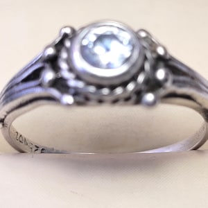 May include: A silver ring with a round, light blue gemstone at the center. The ring has an ornate design with small, raised details around the gemstone and along the band. The band has a split design with engraved lines.