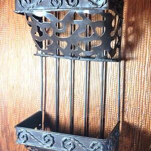 May include: A two-tiered, dark metal wall shelf with decorative scrollwork and cut-out designs. The shelf has two baskets and vertical bars, perfect for displaying items. The metal has a rustic, antique finish, adding a touch of vintage charm.