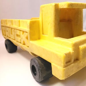 May include: A yellow toy truck with the word "TRUCK" embossed on the side. The truck has black wheels and a simple, blocky design. The toy is made of a solid, possibly plastic, material.