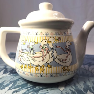 May include: White ceramic teapot with a lid, handle, and spout. The teapot features a whimsical design of cartoon ducks with bows and flowers against a light blue and yellow striped background. Floral and striped patterns adorn the top and bottom.
