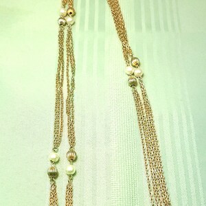 Puede incluir: Un collar dorado con m&uacute;ltiples cadenas y elementos decorativos. El collar presenta peque&ntilde;as perlas redondas y cuentas doradas texturizadas intercaladas a lo largo de las cadenas. El fondo es una tela verde claro.