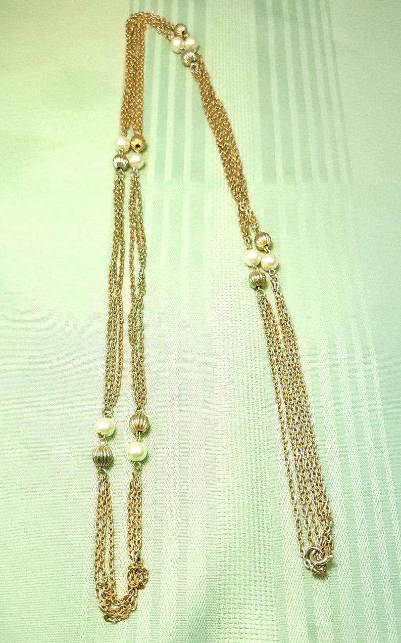 Puede incluir: Un collar dorado con m&uacute;ltiples cadenas y elementos decorativos. El collar presenta peque&ntilde;as perlas redondas y cuentas doradas texturizadas intercaladas a lo largo de las cadenas. El fondo es una tela verde claro.