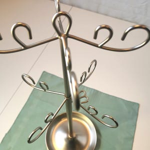 Puede incluir: Un expositor de joyas de metal plateado. El soporte tiene una base circular y múltiples ganchos curvos para colgar collares, pulseras o pendientes. El soporte mide aproximadamente 30 cm de altura.