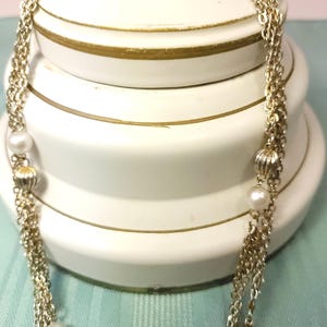 Puede incluir: Un collar dorado con m&uacute;ltiples hilos, con perlas de imitaci&oacute;n y cuentas decorativas acanaladas. El collar est&aacute; drapeado sobre un fondo blanco con ribetes dorados, creando un aspecto cl&aacute;sico y elegante. El collar es un accesorio de moda.