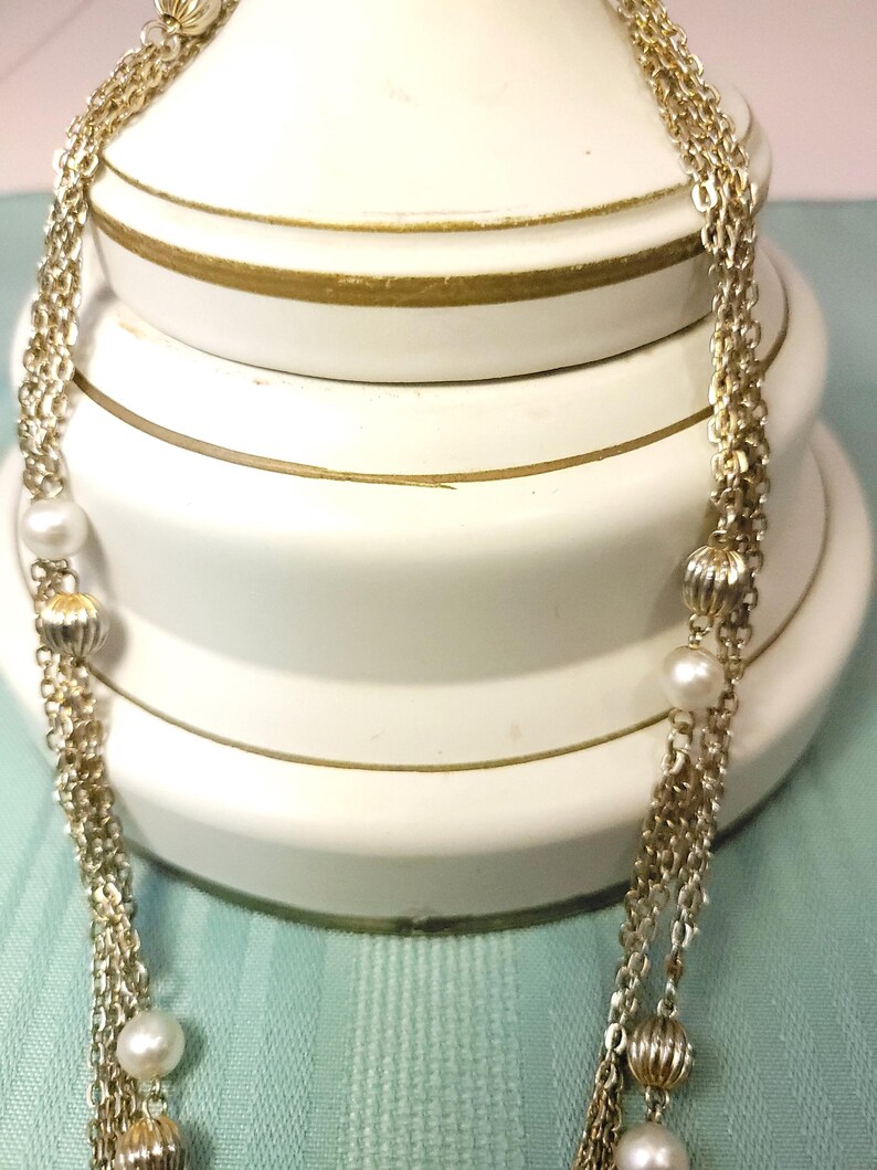Puede incluir: Un collar dorado con m&uacute;ltiples hilos, con perlas de imitaci&oacute;n y cuentas decorativas acanaladas. El collar est&aacute; drapeado sobre un fondo blanco con ribetes dorados, creando un aspecto cl&aacute;sico y elegante. El collar es un accesorio de moda.
