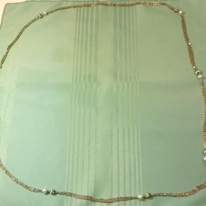 Puede incluir: Un collar largo, dorado, con m&uacute;ltiples hilos y entretejido con peque&ntilde;as perlas blancas redondas. El collar est&aacute; colocado sobre una tela verde claro con rayas verticales.
