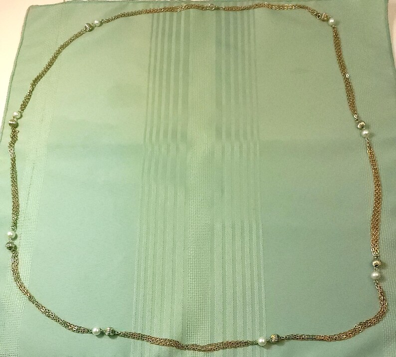 Puede incluir: Un collar largo, dorado, con m&uacute;ltiples hilos y entretejido con peque&ntilde;as perlas blancas redondas. El collar est&aacute; colocado sobre una tela verde claro con rayas verticales.