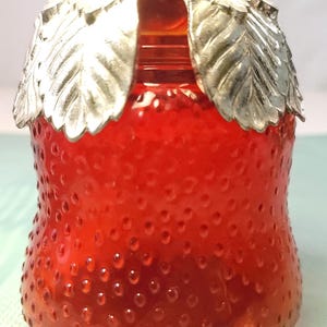 Puede incluir: Un tarro de vidrio rojo con forma de fresa y una tapa plateada con forma de hoja. El tarro tiene una superficie texturizada con pequeñas protuberancias, que se asemejan a una fresa. La tapa tiene un pequeño lazo para facilitar la apertura. Un artículo de cocina decorativo.