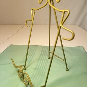 May include: A gold-coloured metal display stand with a three-legged design. The stand features decorative swirls at the base and curved accents at the top, suitable for displaying plates or artwork. The stand is approximately 30 cm tall.