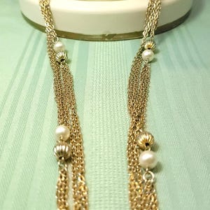 Puede incluir: Un collar dorado con m&uacute;ltiples hilos de cadenas delicadas. El collar presenta perlas de imitaci&oacute;n y cuentas doradas decorativas. La joya se exhibe sobre un fondo verde claro.