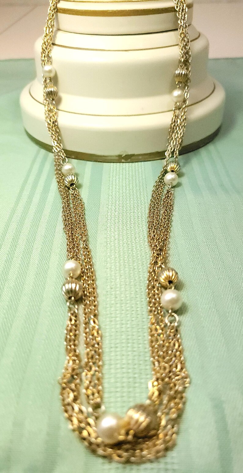 Puede incluir: Un collar dorado con m&uacute;ltiples hilos de cadenas delicadas. El collar presenta perlas de imitaci&oacute;n y cuentas doradas decorativas. La joya se exhibe sobre un fondo verde claro.