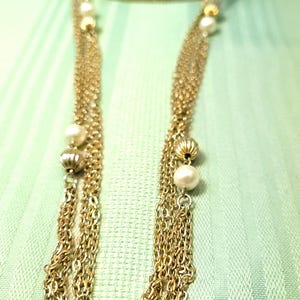 Puede incluir: Collar dorado con m&uacute;ltiples hilos de cadenas finas. El collar presenta perlas de imitaci&oacute;n y cuentas doradas texturizadas. Las cadenas caen en cascada, creando un efecto en capas. El collar se exhibe sobre un fondo verde claro.