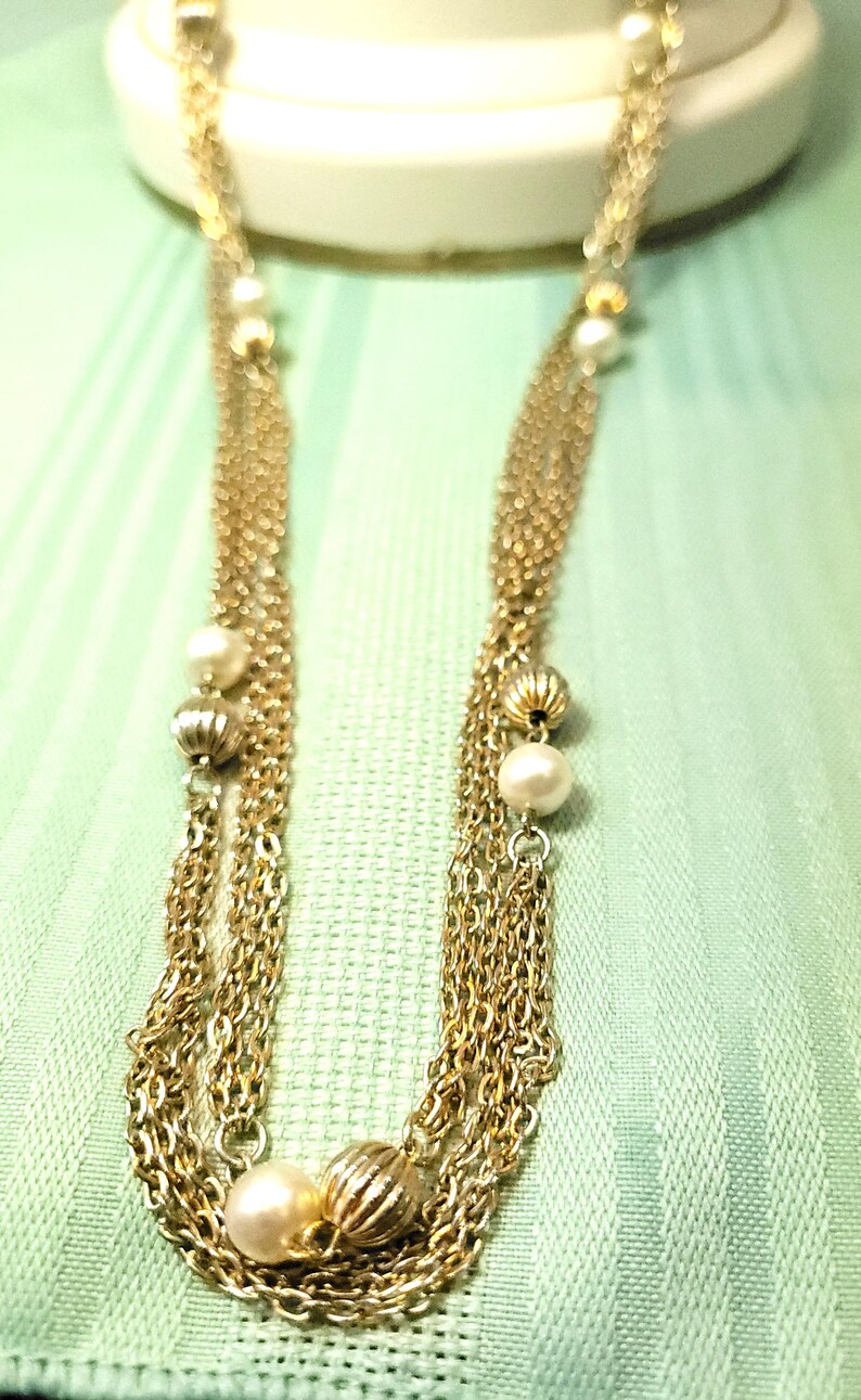 Puede incluir: Collar dorado con m&uacute;ltiples hilos de cadenas finas. El collar presenta perlas de imitaci&oacute;n y cuentas doradas texturizadas. Las cadenas caen en cascada, creando un efecto en capas. El collar se exhibe sobre un fondo verde claro.