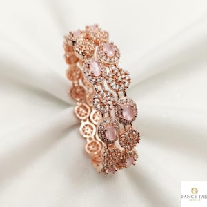 May include: Rose gold bangles with pink oval-shaped gemstones and clear crystals. The jewelry features a repeating pattern of circular and oval designs. The bangles are displayed on a white, textured surface.