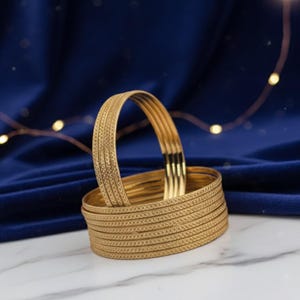 May include: A stack of gold-colored bangles with a textured design. The bangles are arranged on a white marble surface against a dark blue velvet backdrop. The bangles have a repeating pattern and are stacked together.