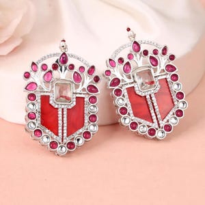 May include: A pair of silver-toned earrings with a shield-like design. Each earring features a red center with a clear, rectangular stone and is surrounded by smaller, pink and clear stones. The earrings have a decorative, ornate design.