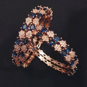 May include: Two rose gold bangles with alternating blue and clear gemstones. The bangles are set against a black background, showcasing the intricate design and sparkle of the jewelry.