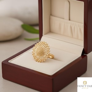 Puede incluir: Un anillo dorado con un diseño de girasol, exhibido en una caja de joyería de madera oscura abierta. El anillo presenta una cabeza y pétalos de girasol detallados y texturizados. La caja de joyería tiene un interior color crema.