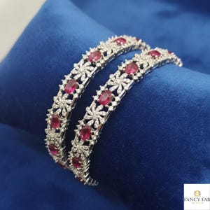 May include: Two silver bangles with a floral design, each featuring oval-shaped red gemstones and small clear stones. The bangles are displayed on a blue velvet surface, showcasing the jewelry's intricate details and sparkle.