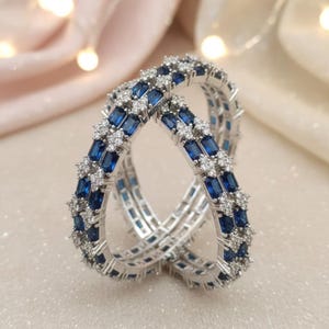 May include: Two silver bangles with alternating rows of blue rectangular and small white round gemstones. The bangles are intertwined, showcasing their intricate design and sparkling appearance. The jewelry is set against a soft, blurred background.