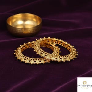 Puede incluir: Dos brazaletes dorados con diseños intrincados se exhiben sobre una superficie de terciopelo morado oscuro. Un pequeño cuenco dorado martillado se encuentra en el fondo. Los brazaletes tienen un patrón repetitivo de arcos y pequeñas cuentas.