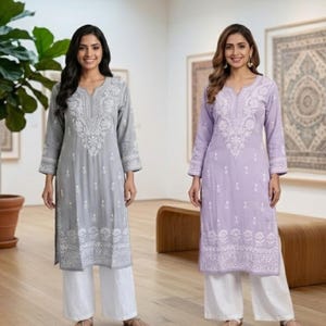 May include: Two long-sleeved tunics featuring intricate white floral embroidery on the chest and hem. One tunic is light gray and the other is light purple. Both are paired with loose-fitting white trousers, creating a coordinated set of garments.