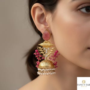 May include: Gold-coloured chandelier earrings featuring a pink gemstone, detailed filigree, and hanging pink beads. The earrings have a bell-shaped base with white pearl accents. The earrings are a statement piece.