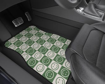 May include: Car floor mat featuring a green and white checkerboard design with smiling faces. The mat is placed in a car, in front of the pedals. It has a black border and a curved shape to fit the car's floor.