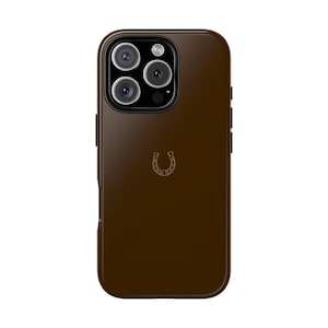 Puede incluir: Una funda de teléfono de cuero marrón con un diseño de herradura dorada. La funda tiene una protuberancia de cámara elevada y un borde negro.