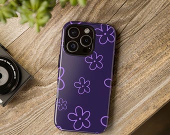 以下が含まれることがあります： 紫色の携帯電話ケース。明るい紫色の花の繰り返し模様が施されています。ケースは木製の表面に置かれています。背景にはカメラと植物が見えます。