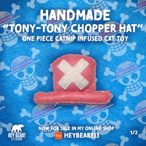 Op de afbeelding: Handgemaakt roze kattenspeelgoed in de vorm van Tony-Tony Choppers hoed, met een wit kruis. De hoed staat op een blauwe achtergrond met een patroon van cartoon schedels en gekruiste botten. Tekst luidt "HANDMADE TONY-TONY CHOPPER HAT" en "ONE PIECE CATNIP INFUSED CAT TOY."