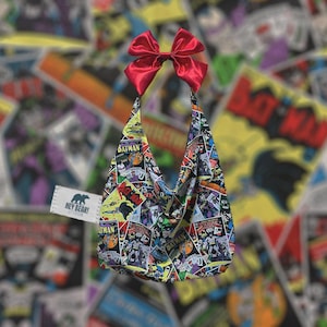 Puede incluir: Bolso de hombro con estampado de cómic de Batman. Cuenta con un lazo de satén rojo en la parte superior y una etiqueta que dice "Hey Bear!". El diseño del bolso incorpora varias portadas de cómics de Batman.