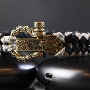 May include: A braided bracelet with a decorative clasp. The bracelet is black, grey, and white. The clasp is gold-coloured with an ornate design. The bracelet is resting on a dark surface.