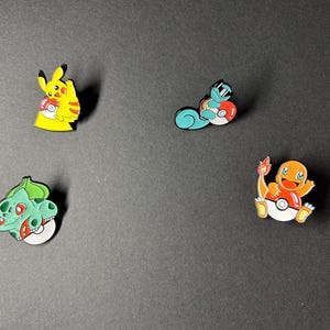 Peut inclure: Quatre épingles en émail colorées représentant des personnages populaires. Pikachu est jaune, tenant une balle rouge et blanche. Carapuce est bleu, avec des lunettes de soleil, et tient une balle rouge et blanche. Bulbizarre est vert, et Salamèche est orange, avec une flamme.