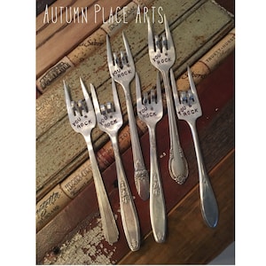 May include: Six silver forks with the words "You Rock" engraved on the tines. The forks are arranged on a wooden surface with cork accents.