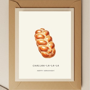 May include: Greeting card featuring a watercolor illustration of a challah loaf with sesame seeds. The card has the text "CHALLAH-LA-LA-LA" and "HAPPY HANUKKAH!" on a cream background, set within a brown envelope.